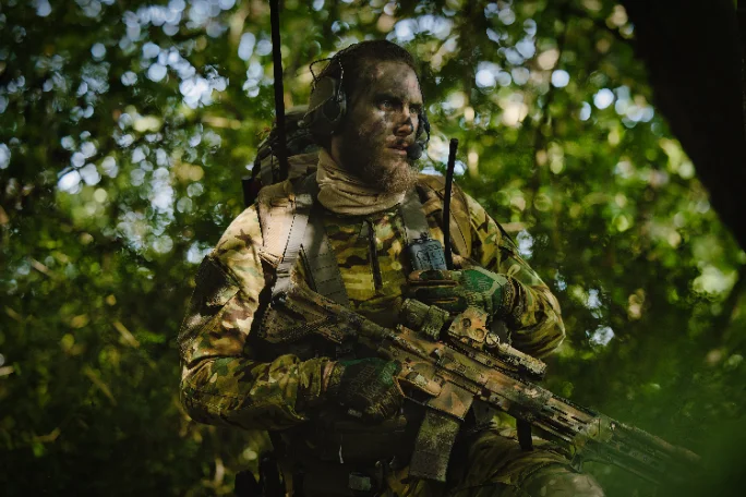 Operator with camouflage face paint and an over ear headset moving through summer woodland with a rifle