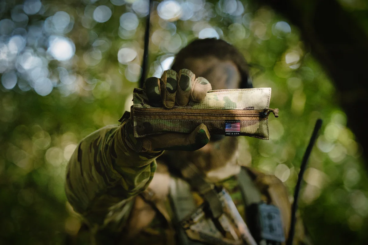 Gloved hand holding out a camouflage pouch toward the camera, operator blurred in the woodland behind