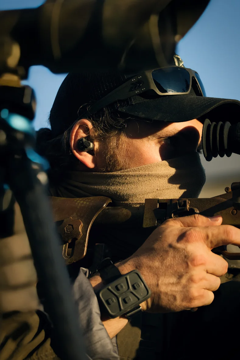 Close view of a marksman behind a scope, hearing protection in his ear, control unit strapped to his wrist
