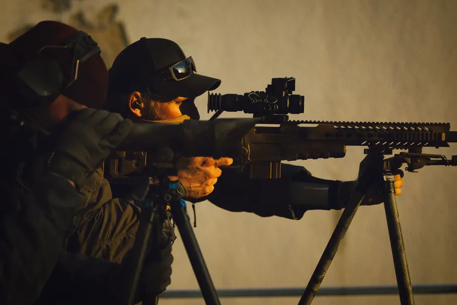 Rifle on a bipod against a wall, shooter behind the optic in low sun
