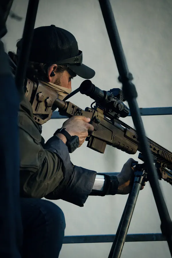 Marksman behind a bolt action rifle on a bipod, cold daylight, scaffold in the frame