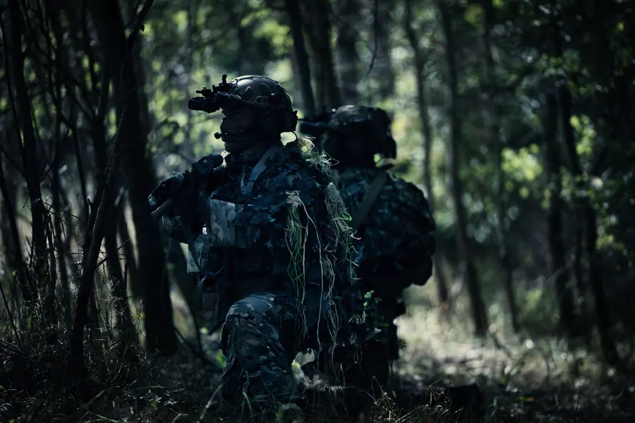 Two operators moving through a dark pine forest, plate carriers and packs silhouetted against the trees