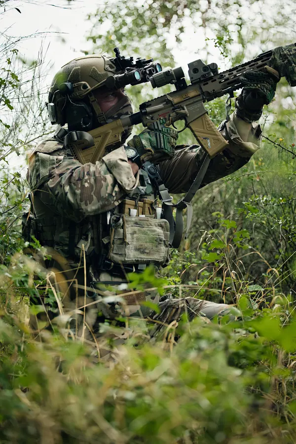 Operator kneeling in tall grass at the treeline, rifle shouldered