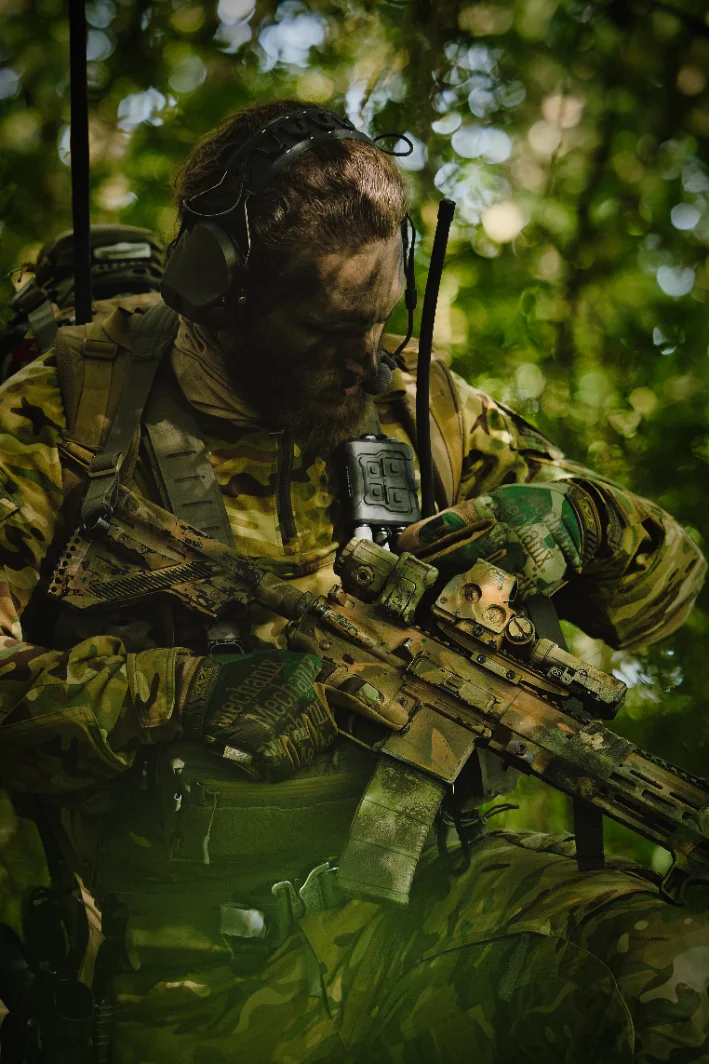 Bearded operator in woodland with an over ear headset and boom microphone, rifle held across his body