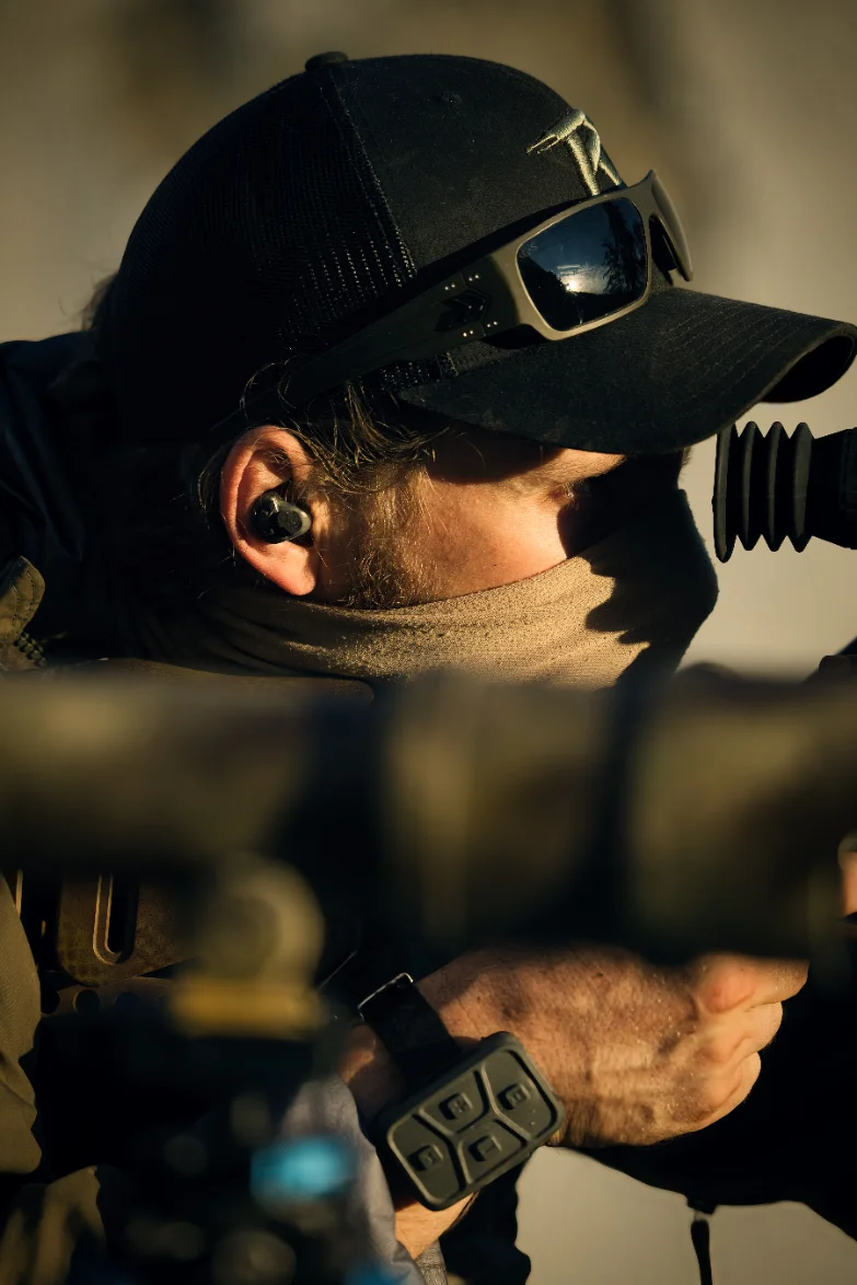 Marksman in a cap and sunglasses behind a scoped rifle, communication earpiece seated in his ear