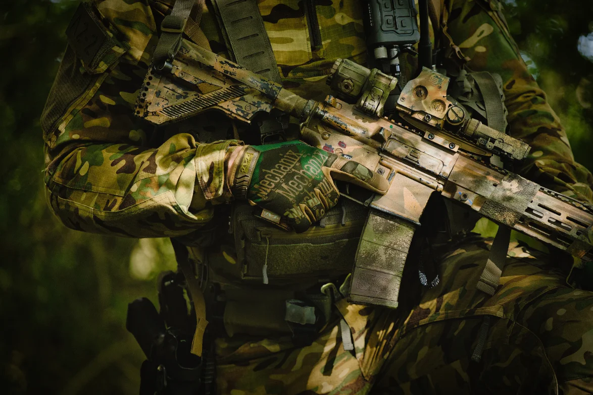 Gloved hand of an operator in woodland camouflage resting on a painted rifle, Mechanix glove logo visible