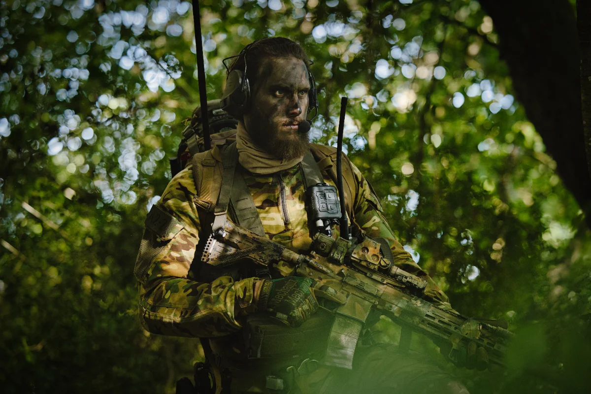 Bearded operator with face paint and a headset standing in dense green woodland, rifle held low