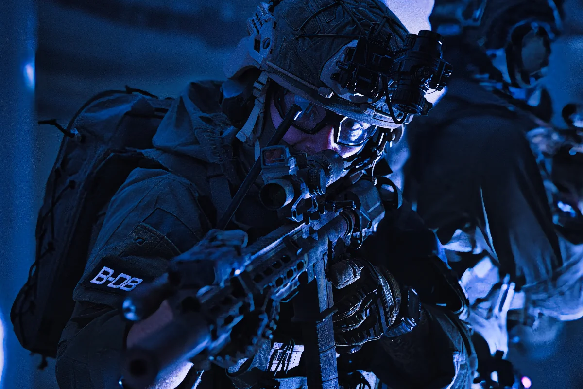 Operator advancing in blue light with a shouldered rifle, gloved support hand on the handguard