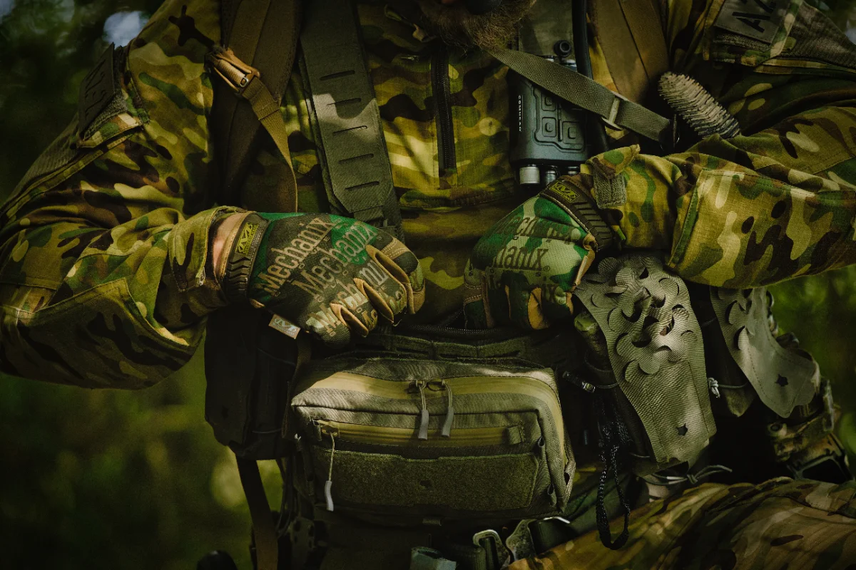 Gloved hands of an operator resting on a chest rig with a waist pouch, camouflage uniform in green woodland