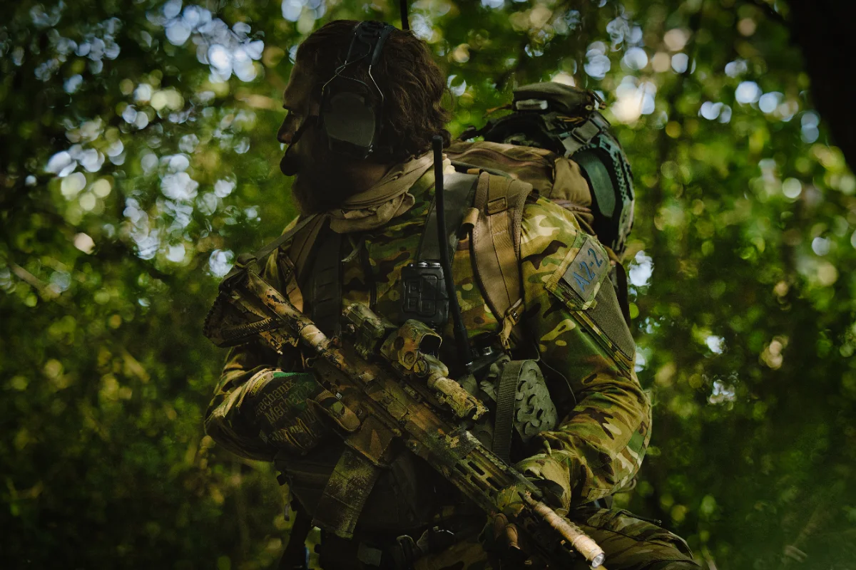 Operator in woodland camouflage looking over his shoulder, headset and radio antenna visible, rifle slung