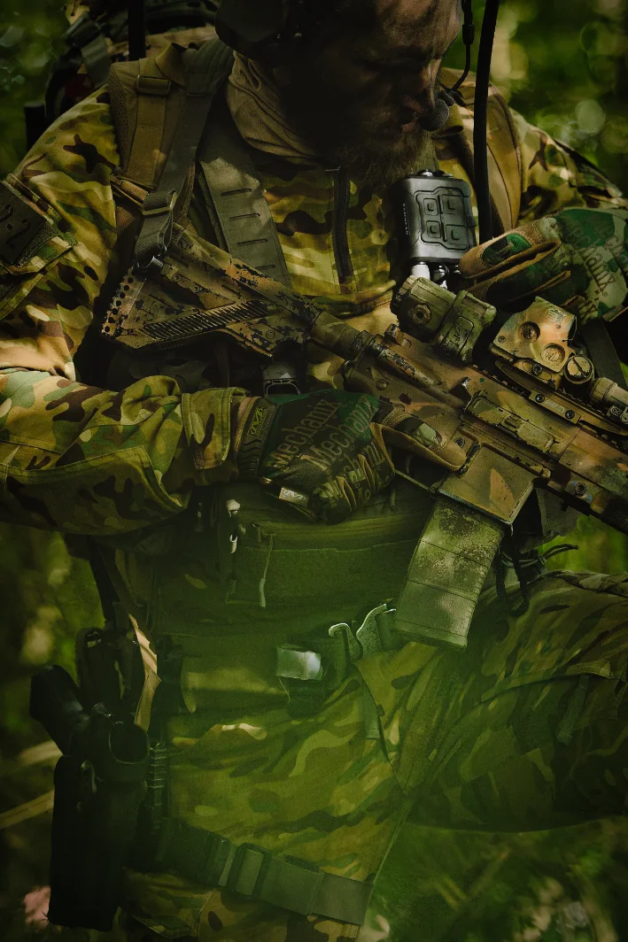 Chest level view of an operator in woodland camouflage, gloved hands on a rifle, radio on the harness